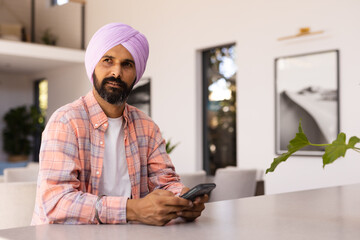 Using smartphone, man in turban sitting at home, looking thoughtful, copy space