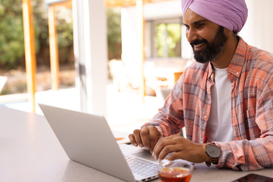 Typing on laptop, smiling man in turban working from home office, copy space