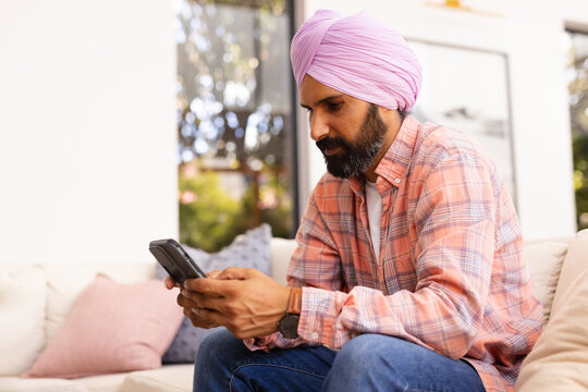 Using smartphone, man in turban sitting on couch at home, concentrating