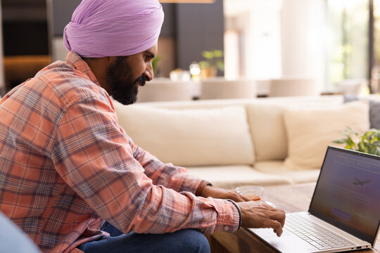 Typing on laptop, man in turban working from home on couch - Powered by Adobe