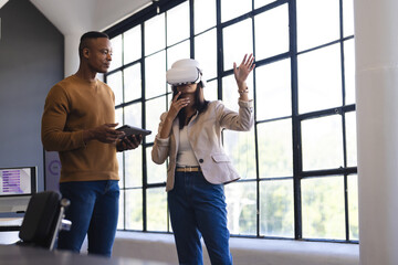 Using VR headset, businesswoman interacting while colleague observes with tablet in office