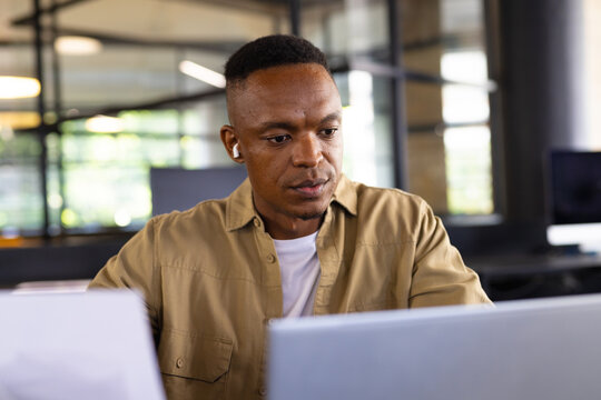 Working in office, man using laptop and wearing wireless earbuds, concentrating