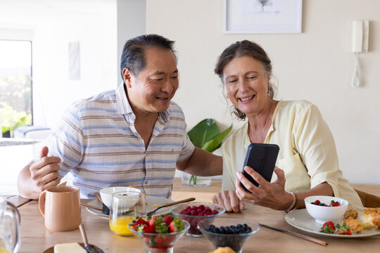Smiling senior couple using smartphone while enjoying breakfast together at home