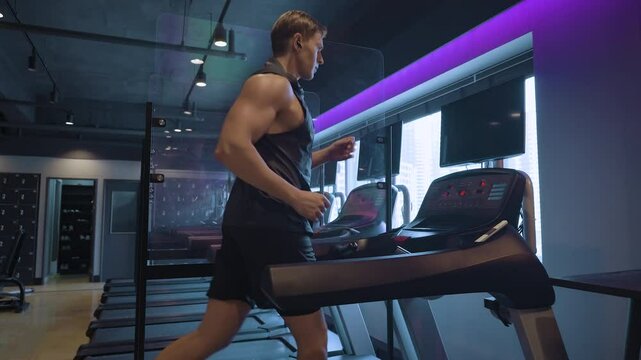 Caucasian Sportsman Jogging on Treadmill Inside Futuristic Health Club in Slow Motion, Listening to Music and Wearing Tank Top Looking At TV Screen - Profile View