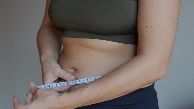 Cropped shot of a young woman in sports clothes measuring a belly girth with a measure tape on a grey background. Close-up. Diet, sport and wellness concept. Excess weight, overweight
