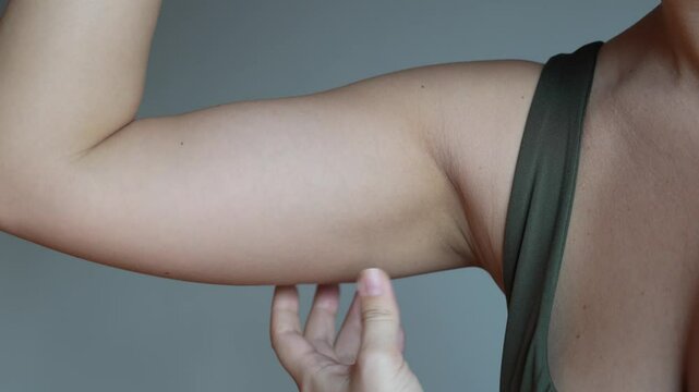 Cropped shot of a young woman grabbing and shaking skin on her upper arm with excess fat on a dark grey background. Pinching the loose and saggy muscles. Overweight concept