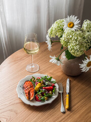 Delicious lunch - grilled salmon, fresh vegetable salad with olives, tomatoes, arugula and a glass of white wine on a wooden table
