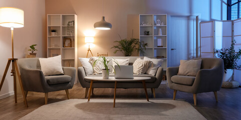 Interior of cozy living room with sofa, table, armchairs and glowing lamps in evening