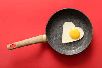 Frying pan with tasty fried egg in shape of heart on red background