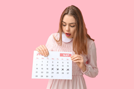 Excited young woman holding calendar with marked days on pink background. Reminder concept