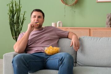 Handsome young man with bowl of tasty potato chips sitting on sofa at home