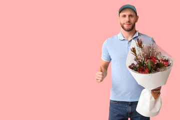 Young delivery man with bouquet of beautiful flowers showing thumb-up gesture on pink background