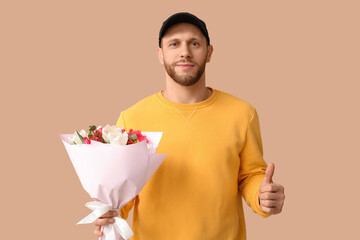 Young delivery man with bouquet of beautiful tulips showing thumb-up gesture on brown background