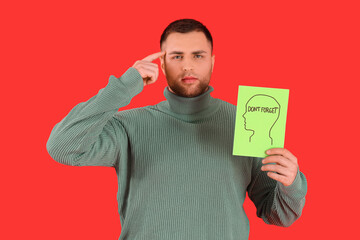 Young man holding paper with text DON'T FORGET and pointing at his head on red background