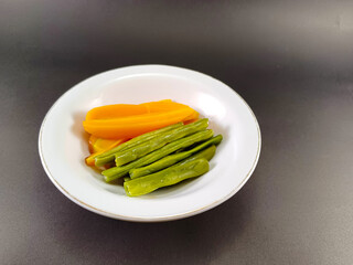 Chopped green beans paired with long sliced orange carrots, artfully arranged in a white bowl, set against a dark grey ash background, creating a vibrant and visually appealing contrast of colors