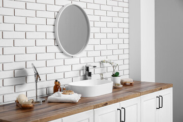 Sink, bottles of cosmetic products on chest of drawers and mirror on white brick wall in bathroom