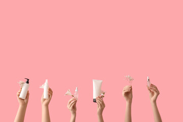 Female hands with bottles of cosmetic products and flowers on pink background