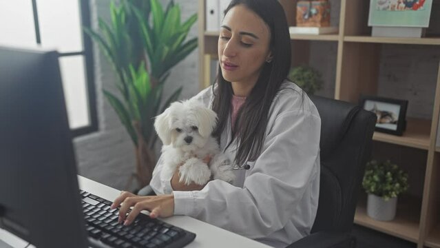 A female veterinarian holding a maltese dog while working at a computer in a vet clinic office.