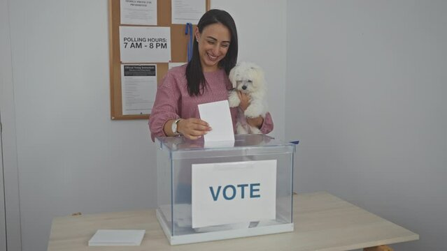 A smiling young woman voting while holding a bichon maltese dog at an indoor electoral college.
