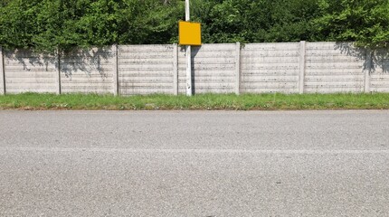 Pole on the roadside with empty yellow signboard on top. Concrete precast compound surrounding wall and trees on behind, strip of grass and road in front. Background for copy space.  