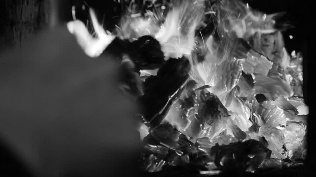 a male hand stirs burning firewood with a poker in the firebox of a stove or fireplace black white video close-up