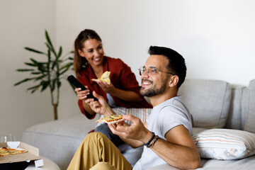 Happy couple watching something funny on TV while eating pizza at home.