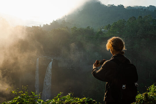 A young girl, a traveler, admires a stunning waterfall and takes pictures with her smartphone.