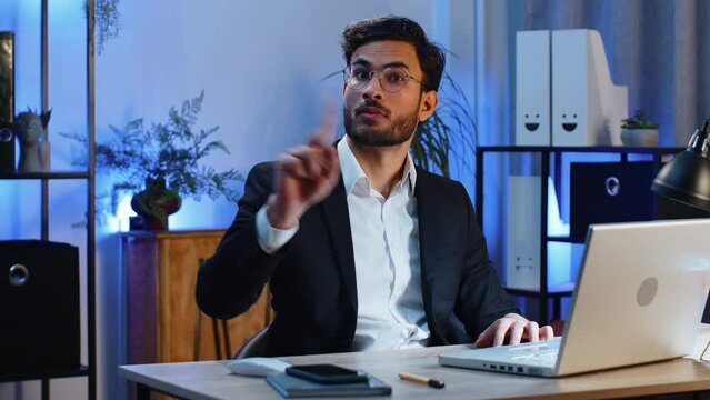 Indian young businessman working on laptop computer shakes finger and saying No be careful scolding and giving advice to avoid danger mistake disapproval sign at home office. Confident freelancer man