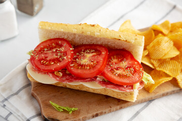 Homemade Italian Sub Sandwich with Salami, Tomatoes and Shredded Iceberg Lettuce on a rustic wooden board, side view.