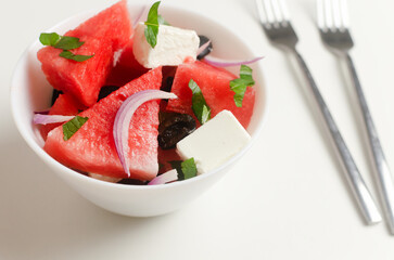 Summer salad with watermelon, feta cheese, olives and mint in a white bowl with two forks on a white table. Concept of vegetarian food. Horizontal orientation.