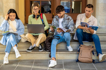 College students with books studying on laptop sitting on staircase of college campus