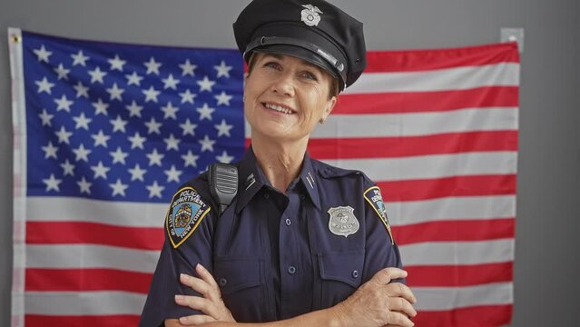 Confident middle-aged woman in police uniform crosses arms in front of american flag, indoors.