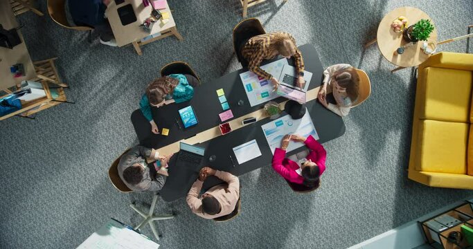 Top Down View: Startup Meeting Room With Team of Diverse Entrepreneurs Sitting at the Conference Table to Discuss Problems, Use Tablet and Laptop Computers, Share Documents with Business Statistics.