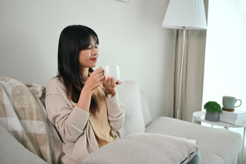 Portrait of attractive Asian woman enjoying coffee while resting on couch in the morning