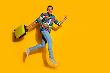Naklejka premium Full size photo of handsome young guy hold suitcase run point empty space wear trendy colorful outfit isolated on yellow color background