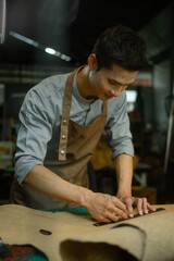 Handsome male tailor in apron measuring and cutting brown leather at his workshop