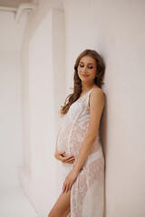 Naklejka premium Beautiful pregnant woman in white retro underwear lace dress and white sandals with high heels. Big pregnant belly. Pretty woman waiting child. Light play on woman's body and face