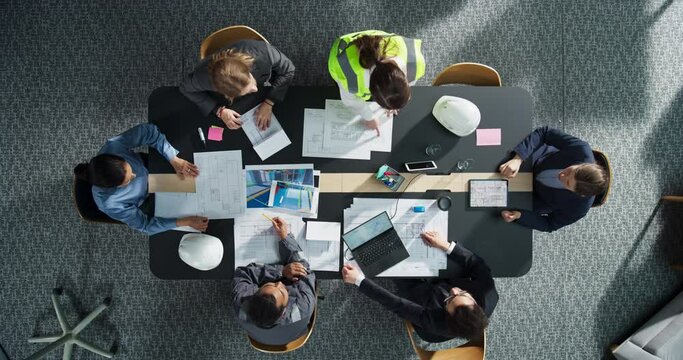 Top Down View of Diverse Team of Real Estate Construction Engineers Discussing Project Blueprints in an Office. Architectural Buro With Colleagues Using Tablet, Laptop, Wearing Reflective Jacket.