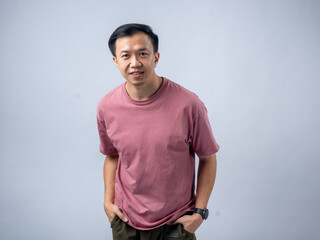 A man in a pink shirt smiles warmly with his hands in his pockets, exuding a relaxed and friendly demeanor. He stands against a plain light blue background,