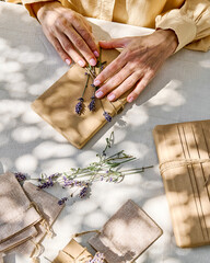Woman making hand made gift package with craft recycled paper and dried lavender flowers on the table with linen tablecloth. Natural aesthetic.