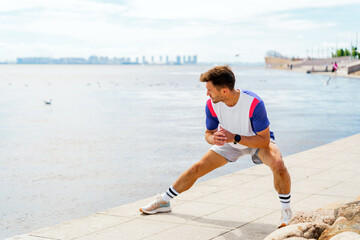 Athletic man stretches by the waterfront, engaging in a dynamic warm-up amidst serene scenery.