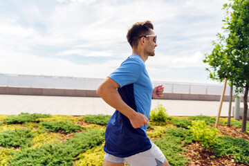 Man athlete in blue sportswear, jogging along a waterfront promenade surrounded by greenery under a...