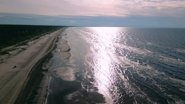 A backward aerial drone shot over the Baltic coast, showcasing the beach with gentle sea waves and sunlight reflecting on the water, creating a beautiful and tranquil scene. Palanga, Lithuania