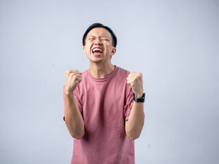 Fototapeta premium A man in a pink shirt raises both fists in a celebratory gesture, his face beaming with excitement and joy. The plain light blue background emphasizes his triumphant and enthusiastic expression.