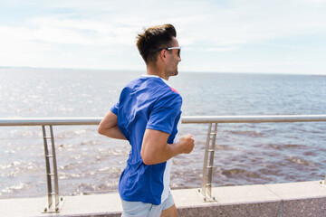 Runner man athlete in tricolor sportswear, jogging along a waterfront promenade under a bright, clear sky.