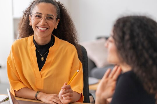 A smiling woman with glasses holds a pen during a meeting, interacting with her colleagues in a bright, modern office environment