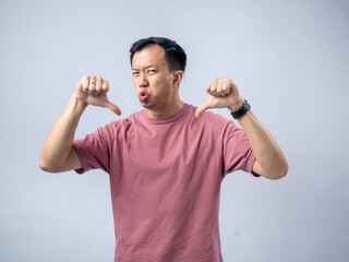 A man in a pink shirt shows a thumbs-down gesture with a displeased expression, sticking out his...