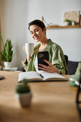 A woman sits at her desk, smiling as she drinks coffee and looks at her phone.