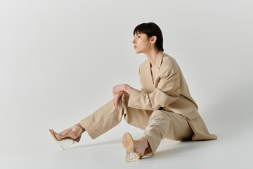 A woman in a tan suit sits on a white background, wearing clear heels.