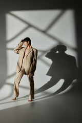 A woman in a beige suit poses against a white backdrop with geometric shadows.
